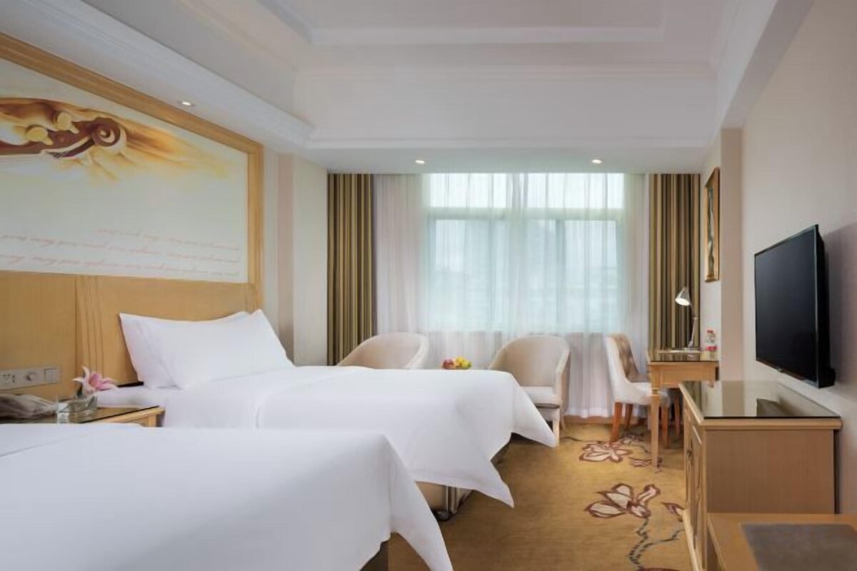 A hotel room with two beds, a television, a desk, and a large window with curtains.