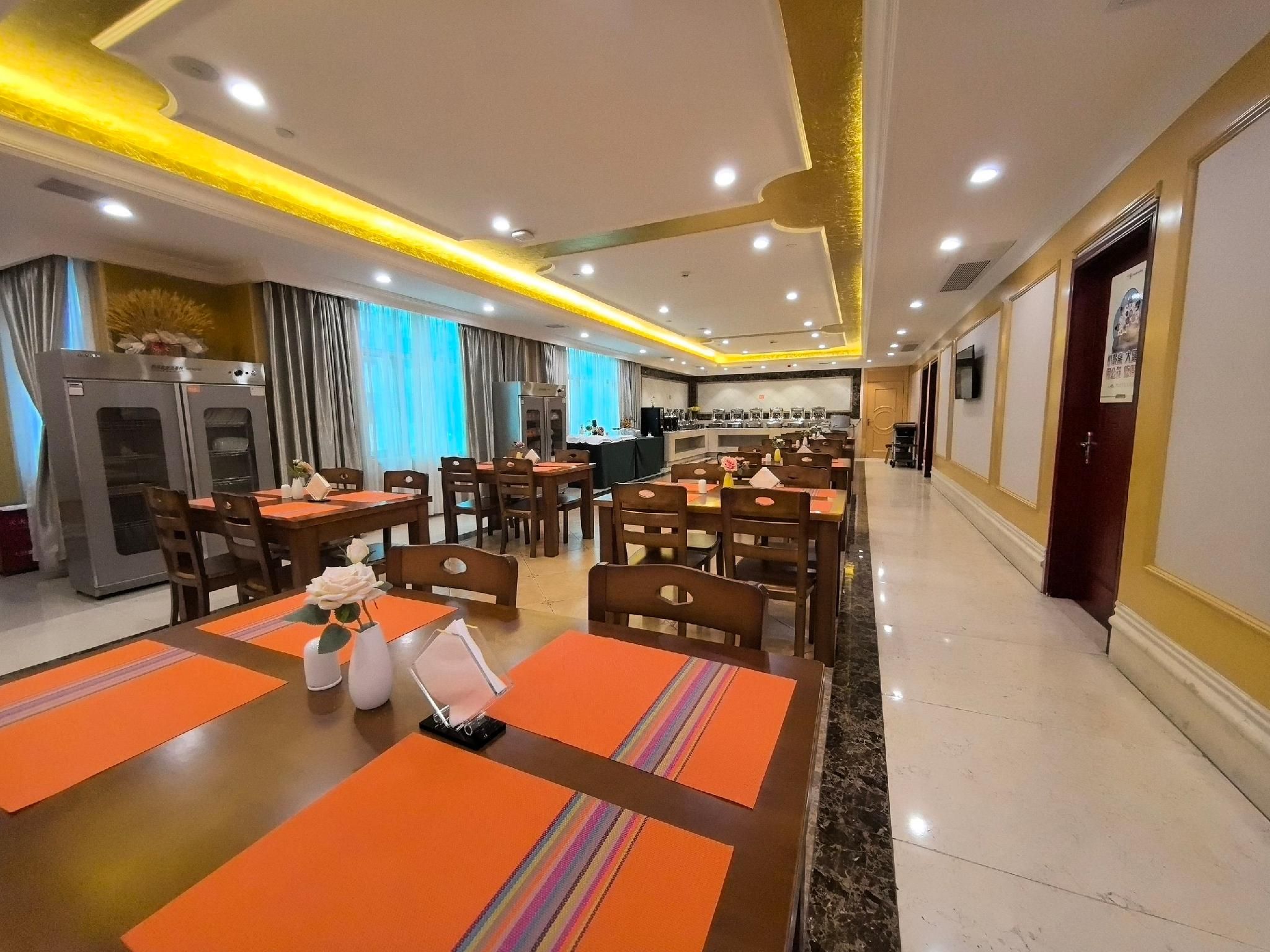 A hotel dining area with multiple tables and chairs, a kitchen in the background, and a modern ceiling design.