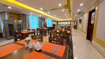 A hotel dining area with multiple tables and chairs, a kitchen in the background, and a modern ceiling design.