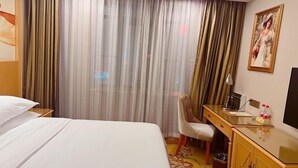 A hotel room with a bed, a desk, a chair, a trash bin, and a painting on the wall.