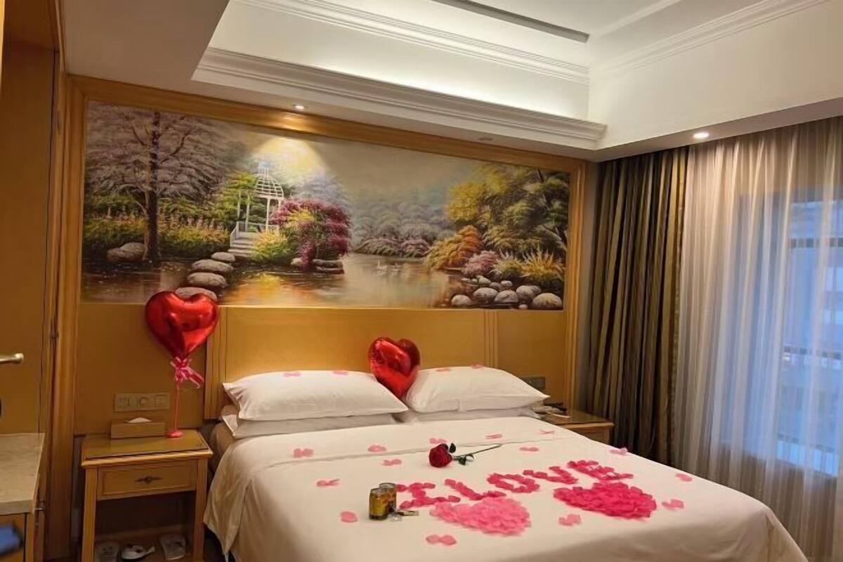A hotel room with a bed, a nightstand, a painting of a garden, and a window with curtains.