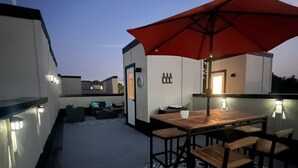 Eine Terrasse im Freien mit einem Holztisch und -stühlen, einem roten Sonnenschirm und einer Bar mit einem Schild.