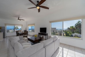A modern living room with a large sofa, a dining table, and a TV. There are large windows offering a view of the sea and palm trees.