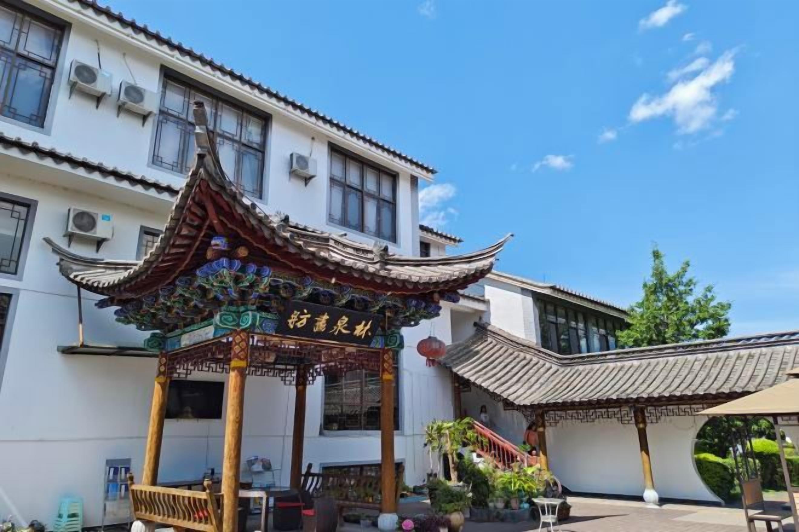 A traditional Chinese pavilion with ornate roof and columns, situated in front of a building with multiple windows and a tiled roof.