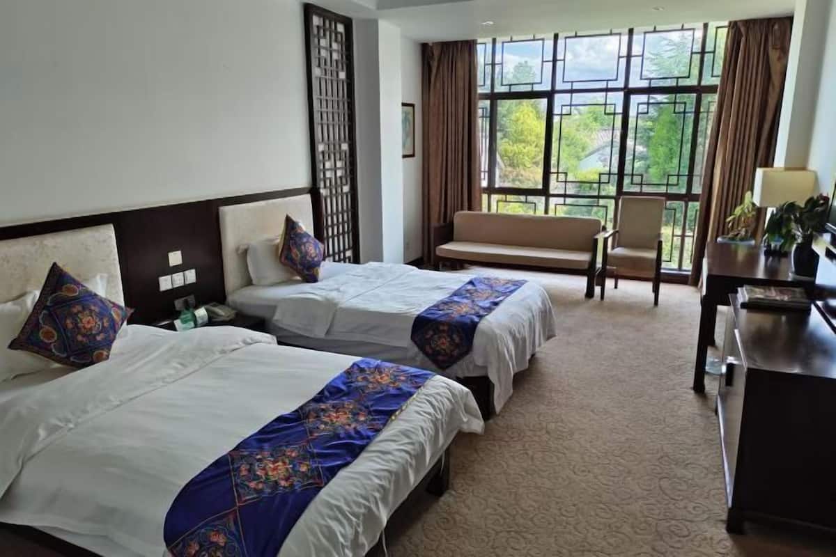 Ein Hotelzimmer mit zwei Betten, einem Sofa, einem Schreibtisch und großen Fenstern mit Blick auf Grünflächen.