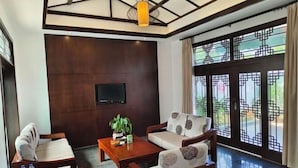 Une chambre d’hôtel avec un plafond en bois, un téléviseur fixé au mur, un canapé, une table basse et une grande fenêtre ornée d’un motif en treillis décoratif.