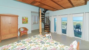 A bedroom with a bed, a wooden wardrobe, a chair, a painting, a spiral staircase, and a balcony with a view. - Bean Point Beachfront- Beachfront 4 bedroom private home with pool/spa (Anna Maria)