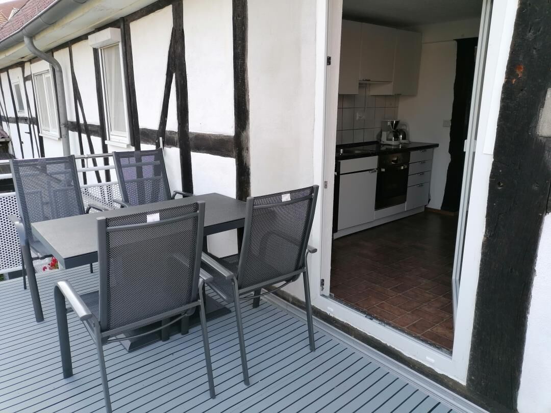 Un comedor con mesa y sillas, una cocina visible a través de un umbral abierto, y un balcón con barandilla.