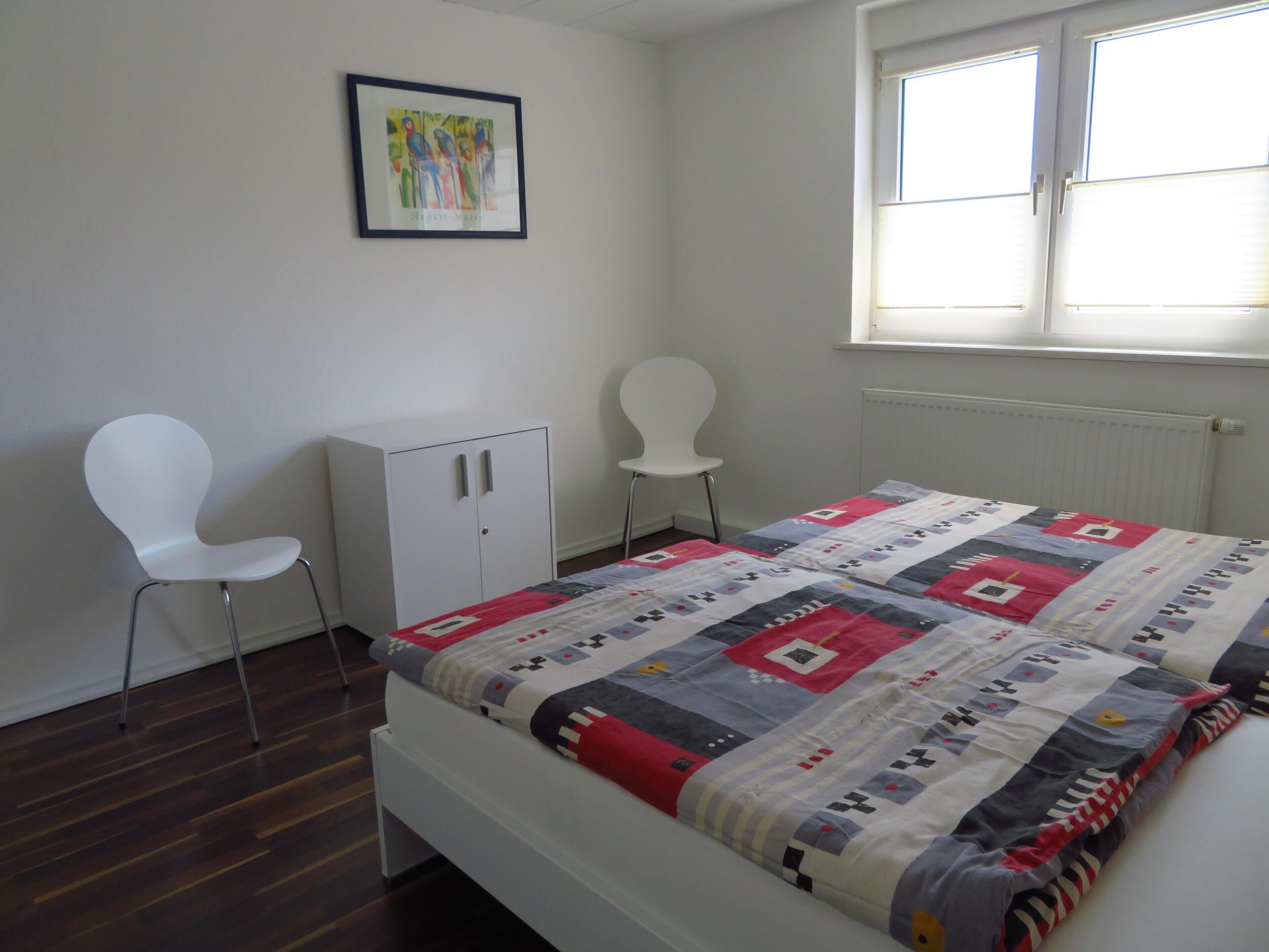 Een slaapkamer met een bed, twee witte stoelen, een witte kledingkast en een raam met zonwering.