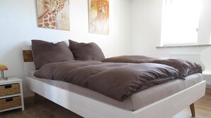 A modern bedroom with a white bed, brown bedding, and wooden flooring. There are two bedside tables with baskets and a lamp. - Apartment Oels Witzenhausen OG incl. balcony (Witzenhausen)