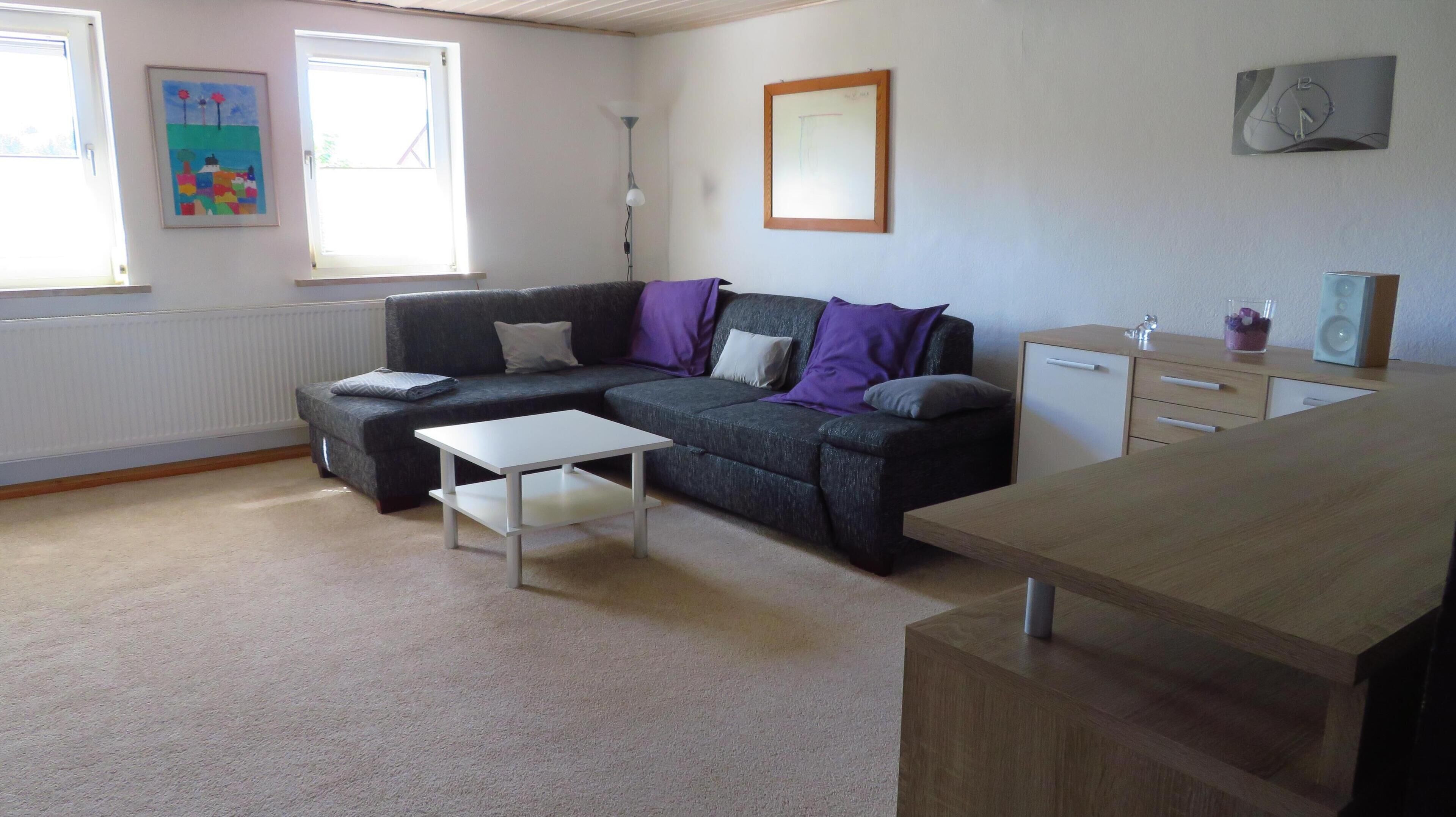 A living room with a grey sofa, a white coffee table, a wooden desk, and a radiator. There are two windows, a painting, and a framed picture on the wall.