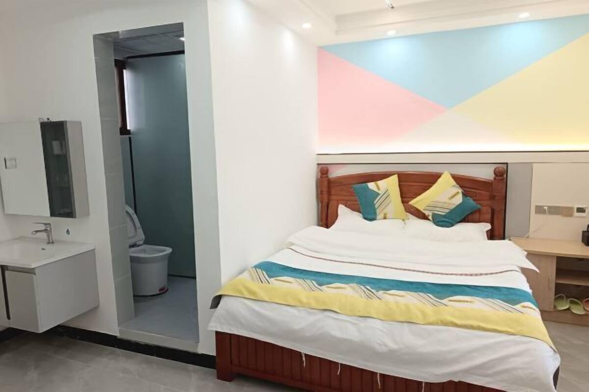 A bedroom with a bed, a bedside table, a mirror, and a bathroom visible through an open door.