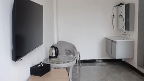 Une chambre moderne équipée d’un téléviseur à écran plat, d’une petite table ronde avec une bouilloire, d’une chaise en osier, d’un lavabo blanc et d’un meuble avec un miroir.