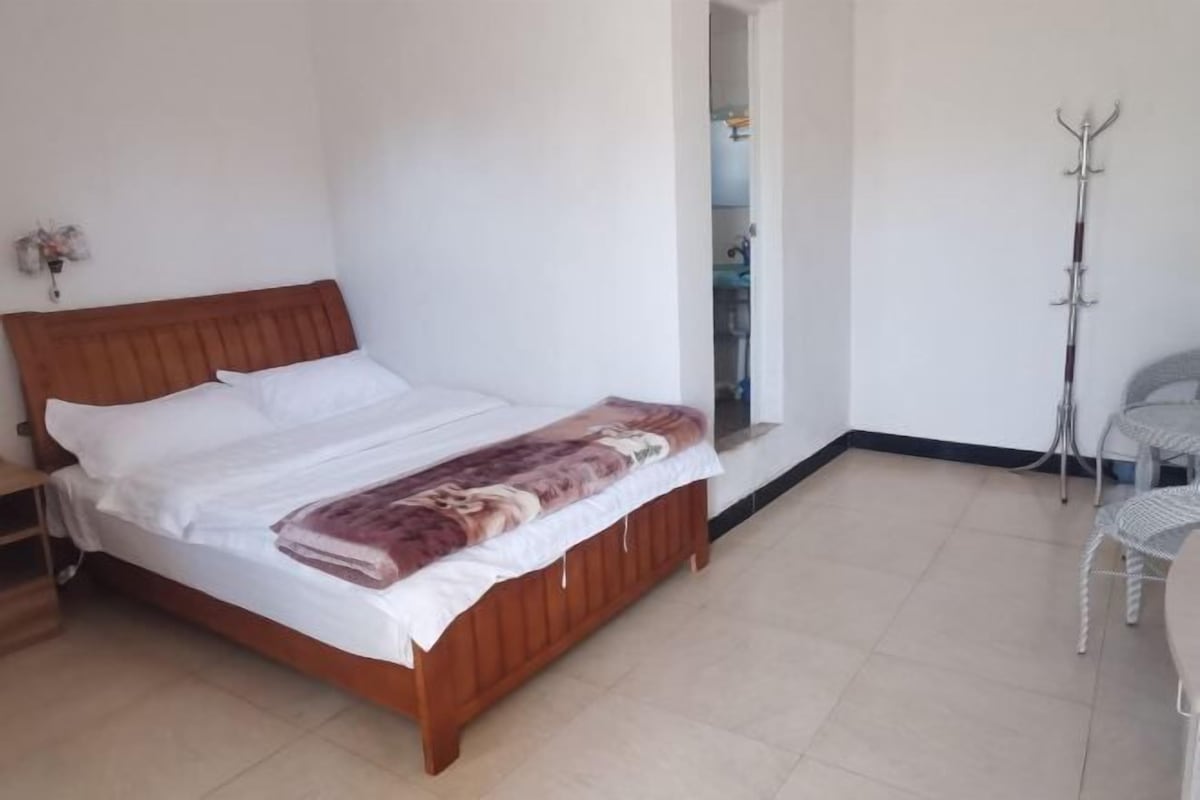 A bedroom with a wooden bed, white bedding, and a patterned throw. There is a chair and a coat rack in the corner.