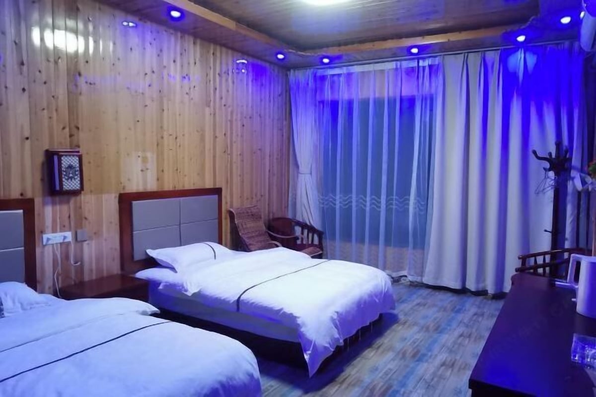A room with wooden walls, two beds with white bedding, a chair, a table, and a window with curtains.