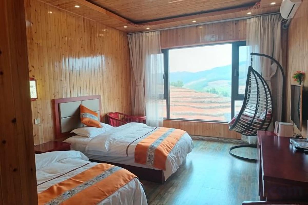 A room with two beds, a hanging chair, a desk with a computer, and a large window with a view of the countryside.