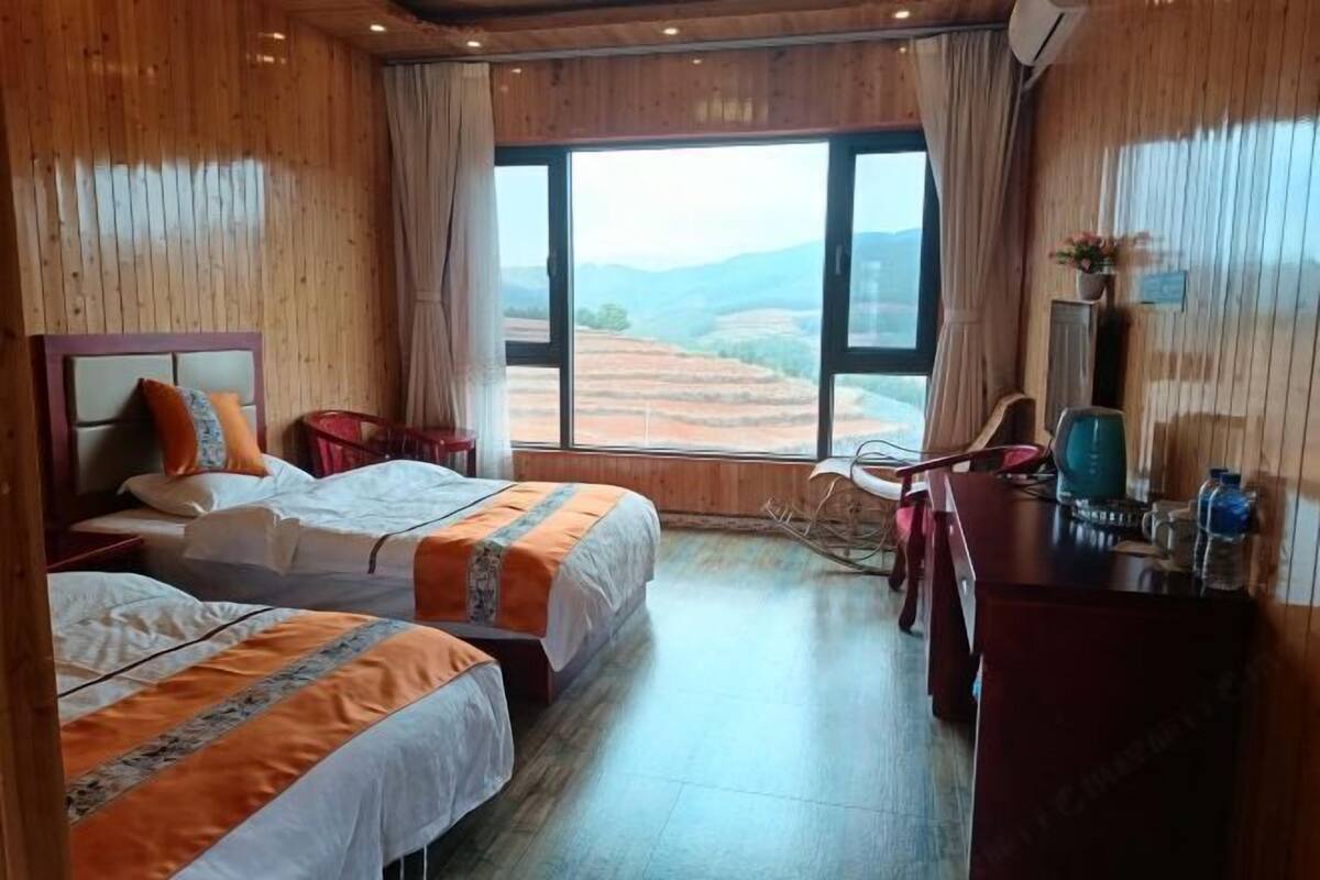 A hotel room with two beds, a desk, a chair, a large window with a view of terraced fields, and a wooden interior.