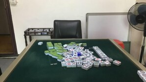 A room with a green felt table set up for playing mahjong. There are multiple sets of mahjong tiles on the table, and a black office chair is visible in the background.