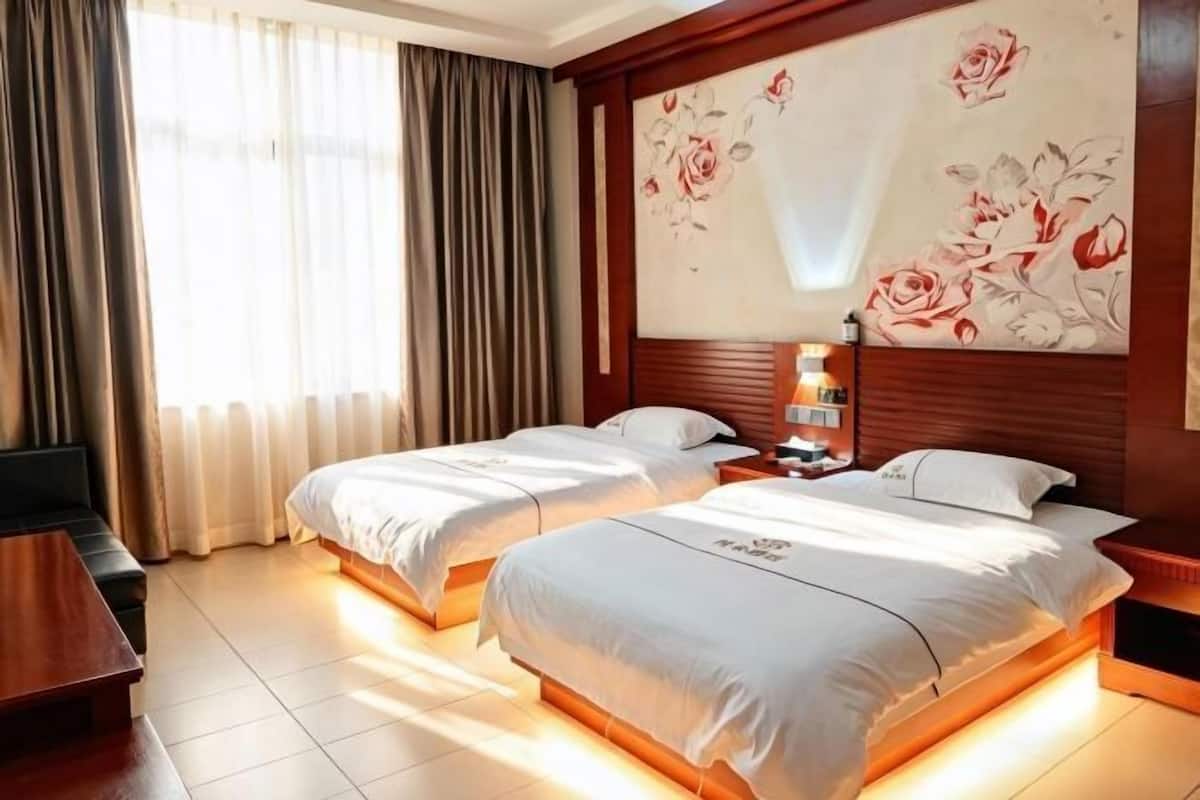 Habitación de hotel con dos camas, cabecero de madera, una obra de arte floral grande, una mesita de noche con una lámpara y vistas al exterior a través de cortinas.