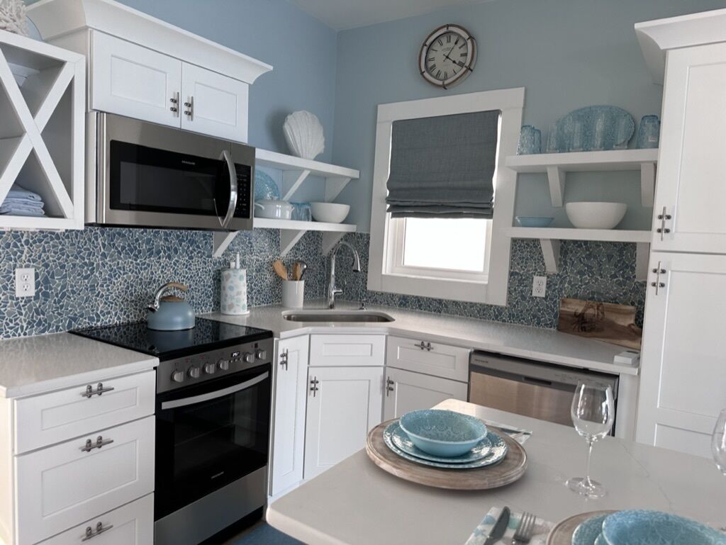 A modern kitchen with white cabinetry, a stainless steel microwave, an oven, and a sink. There is a window with a blind, and a clock on the wall.