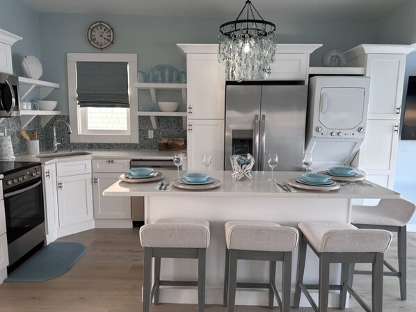 A modern kitchen with white cabinets, a central island with bar stools, a stainless steel refrigerator, and a chandelier.