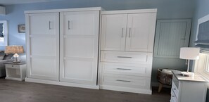 A room with a large white wardrobe, a small bedside table with a lamp, and a wicker basket.