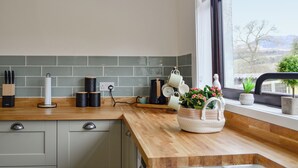 Une cuisine avec un plan de travail en bois, des placards gris, une fenêtre donnant sur les montagnes et un panier de fleurs posé sur le rebord de la fenêtre.