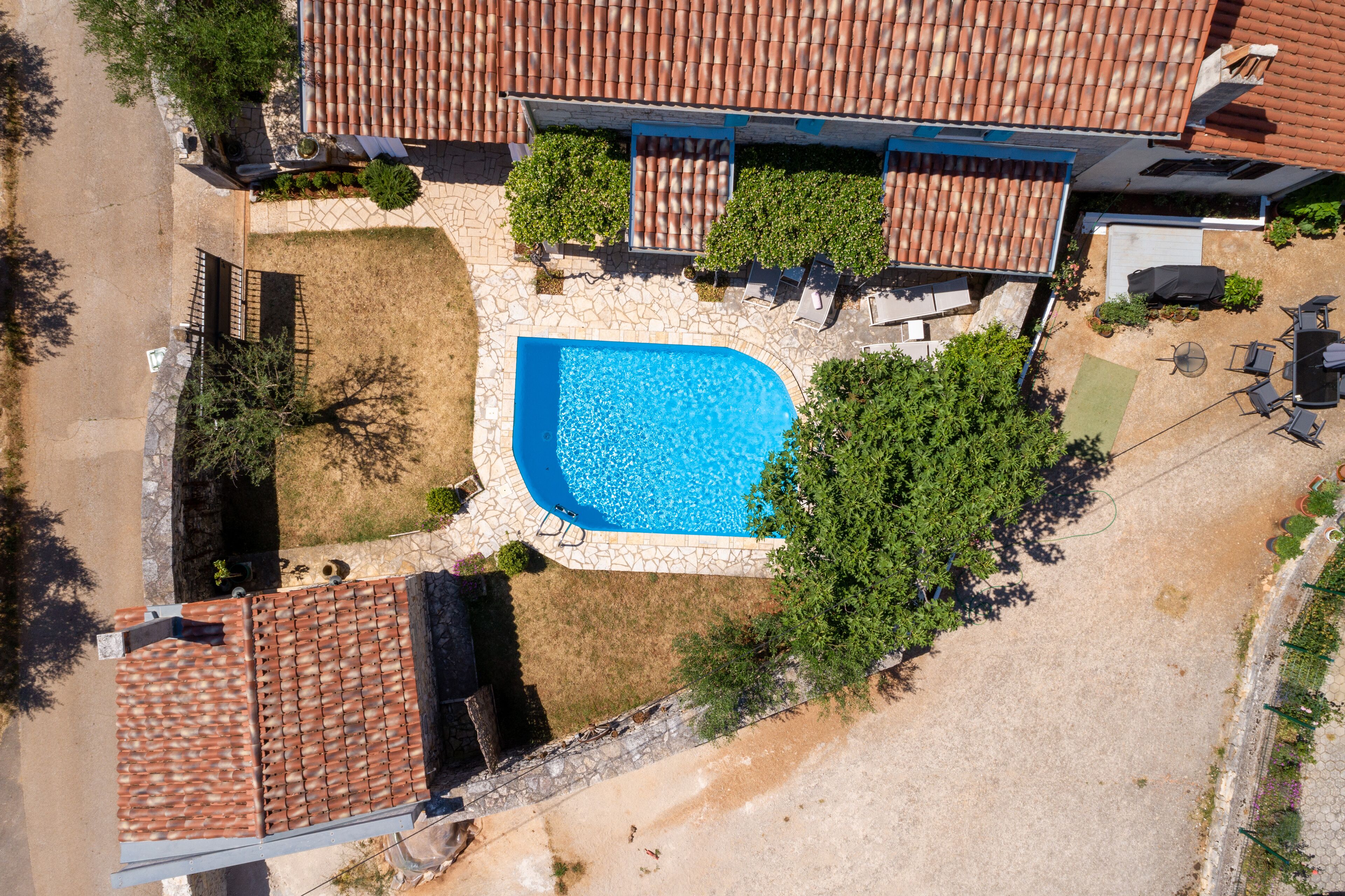 An aerial view of a property with a swimming pool surrounded by a stone patio, a covered outdoor seating area, and a garden with various plants.