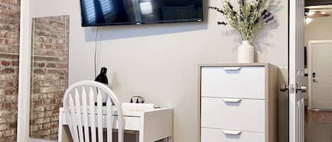 Een kamer met een wit bureau, een stoel, een aan de muur bevestigde televisie, een kast met lades, een potplant en een bakstenen muur.