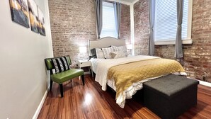 Un dormitorio con pared de ladrillo, una cama con cabecera, un banco y un pequeño otoman.