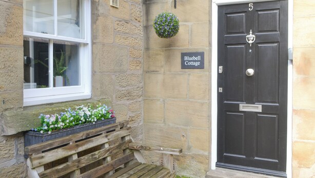 A stone building with a black door marked '5', a wooden bench, and a flower box with white and pink flowers. - 1 bedroom accommodation in Alnwick (Alnwick)