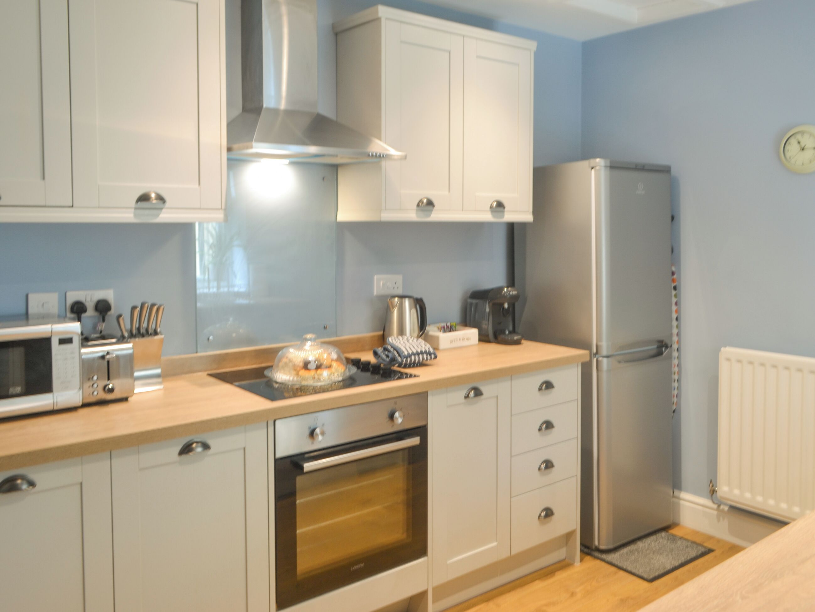 A modern kitchen with white cabinets, a stainless steel range hood, a built-in oven, a microwave, a refrigerator, and a coffee maker.