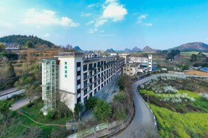 Un edificio de varios pisos con fachada moderna de vidrio, rodeado por un paisaje rural con montañas al fondo.