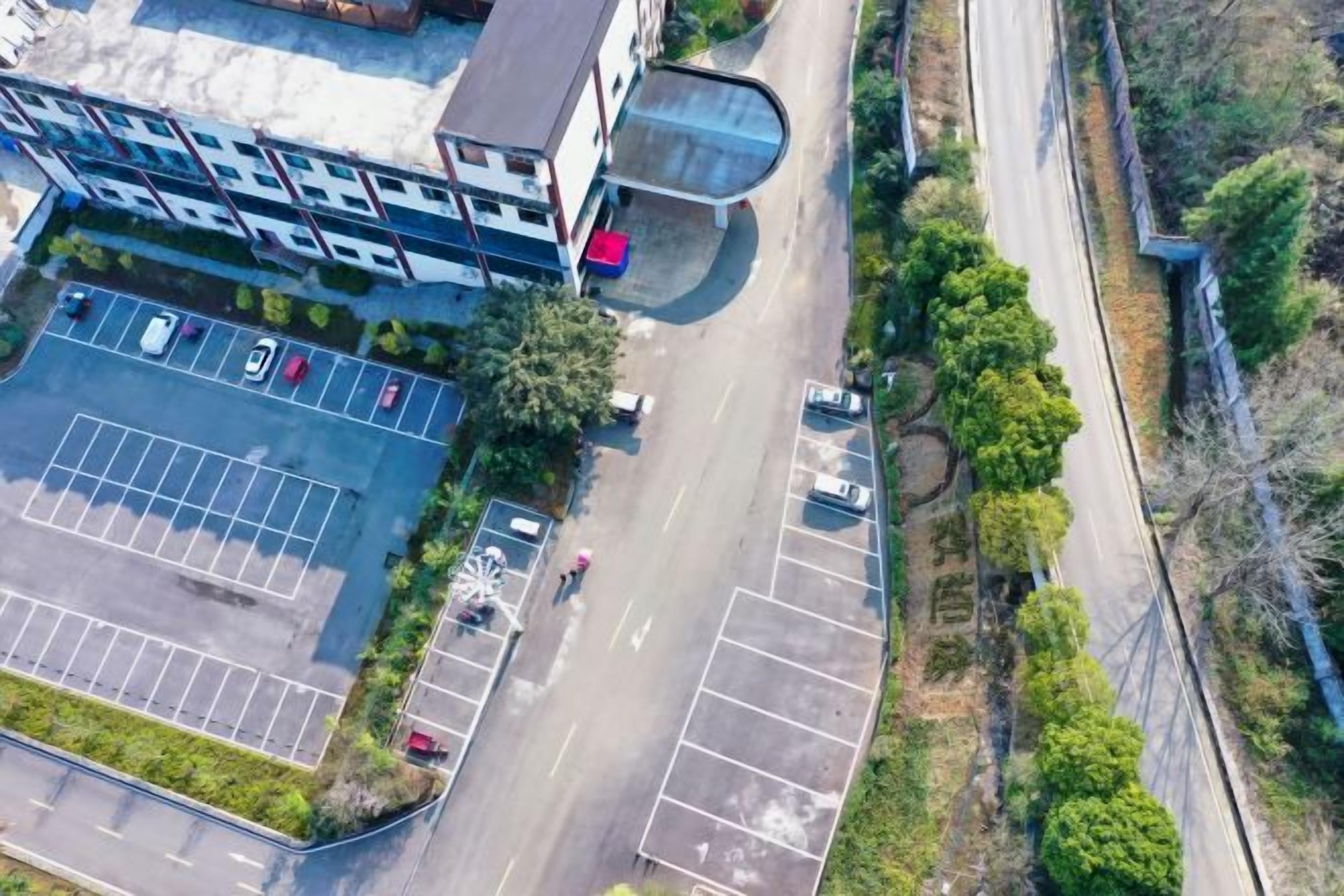 Vista aérea de un estacionamiento con múltiples filas de espacios, un edificio con fachada de vidrio curvo y una carretera adyacente al área de estacionamiento.