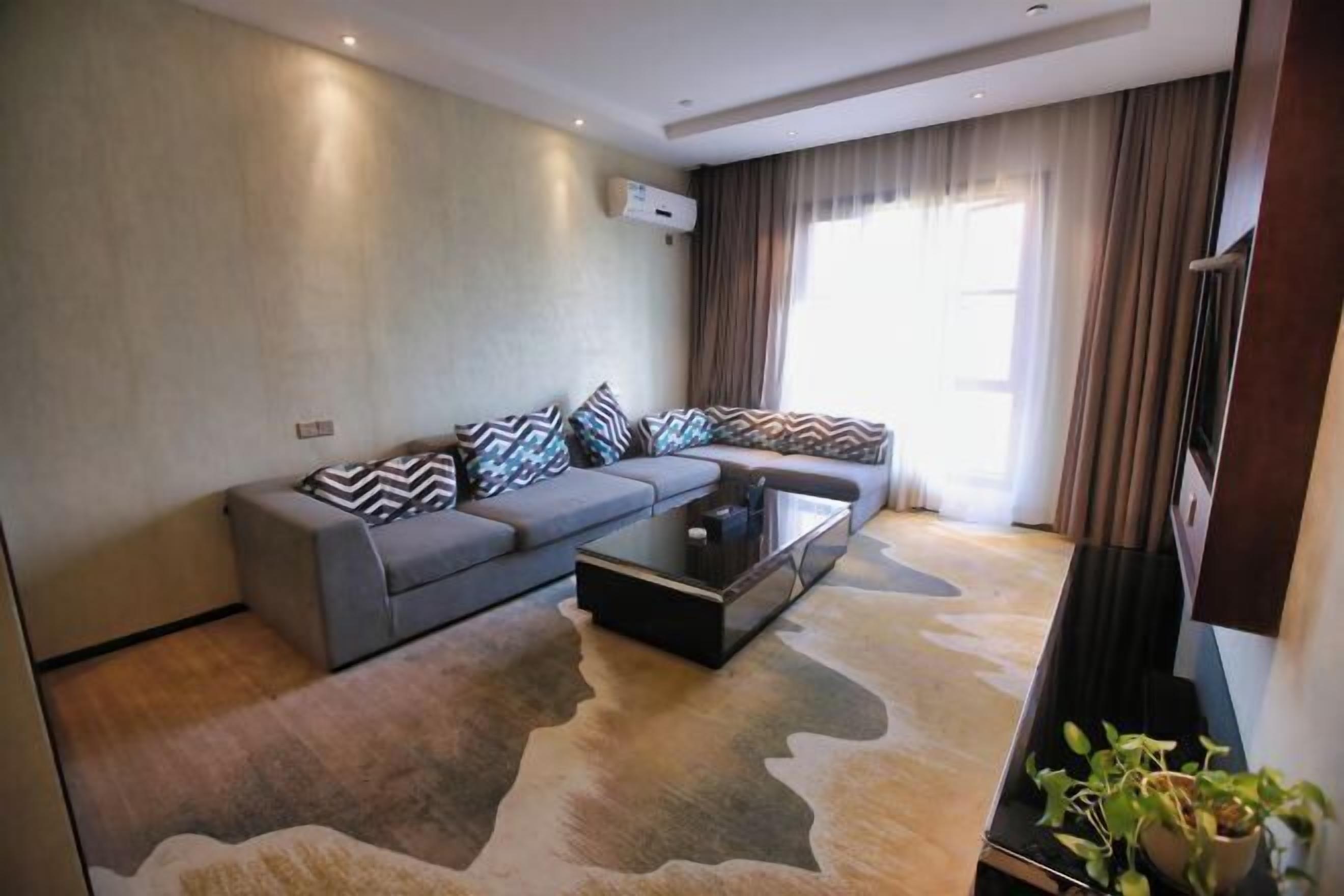A modern living room with a grey sofa, patterned cushions, a glass coffee table, and a large window with curtains.