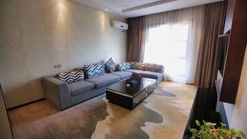 A modern living room with a grey sofa, patterned cushions, a glass coffee table, and a large window with curtains.