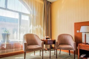 Habitación de hotel con dos sillones, una mesita redonda con lámpara y vista a un edificio a través de un ventanal grande.