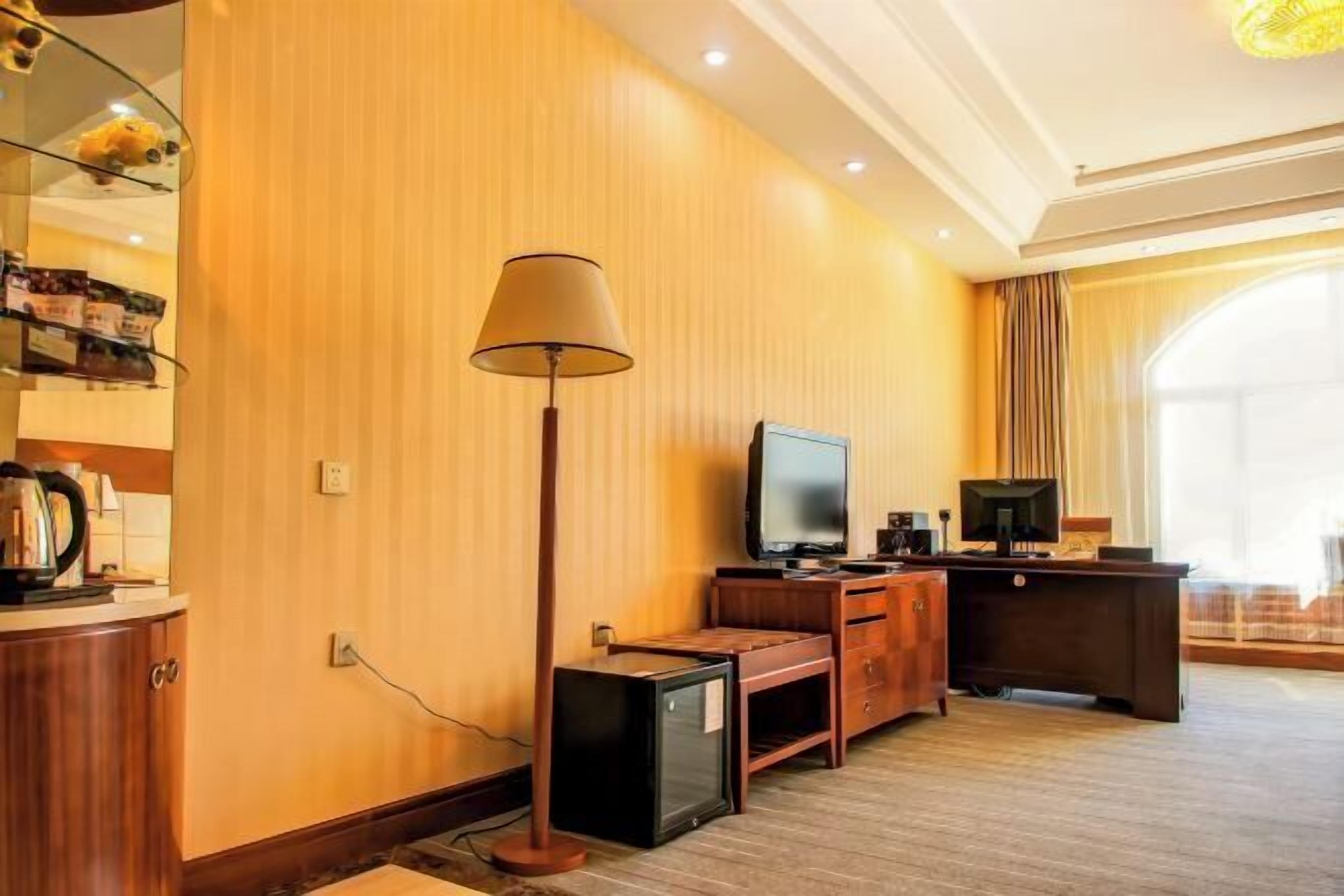 A hotel room with a wooden wall, a flat-screen TV, a desk, a lamp, and a window with curtains.