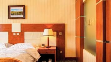 A hotel room with a wooden headboard, a bedside table with a lamp, a framed picture on the wall, and a shoe rack.