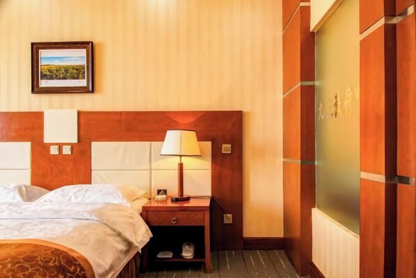 A hotel room with a wooden headboard, a bedside table with a lamp, a framed picture on the wall, and a shoe rack.