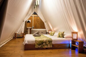 A cozy, tent-style bedroom with a wooden floor, a bed with a green leaf-patterned bedspread, and a small wooden nightstand with a lamp.