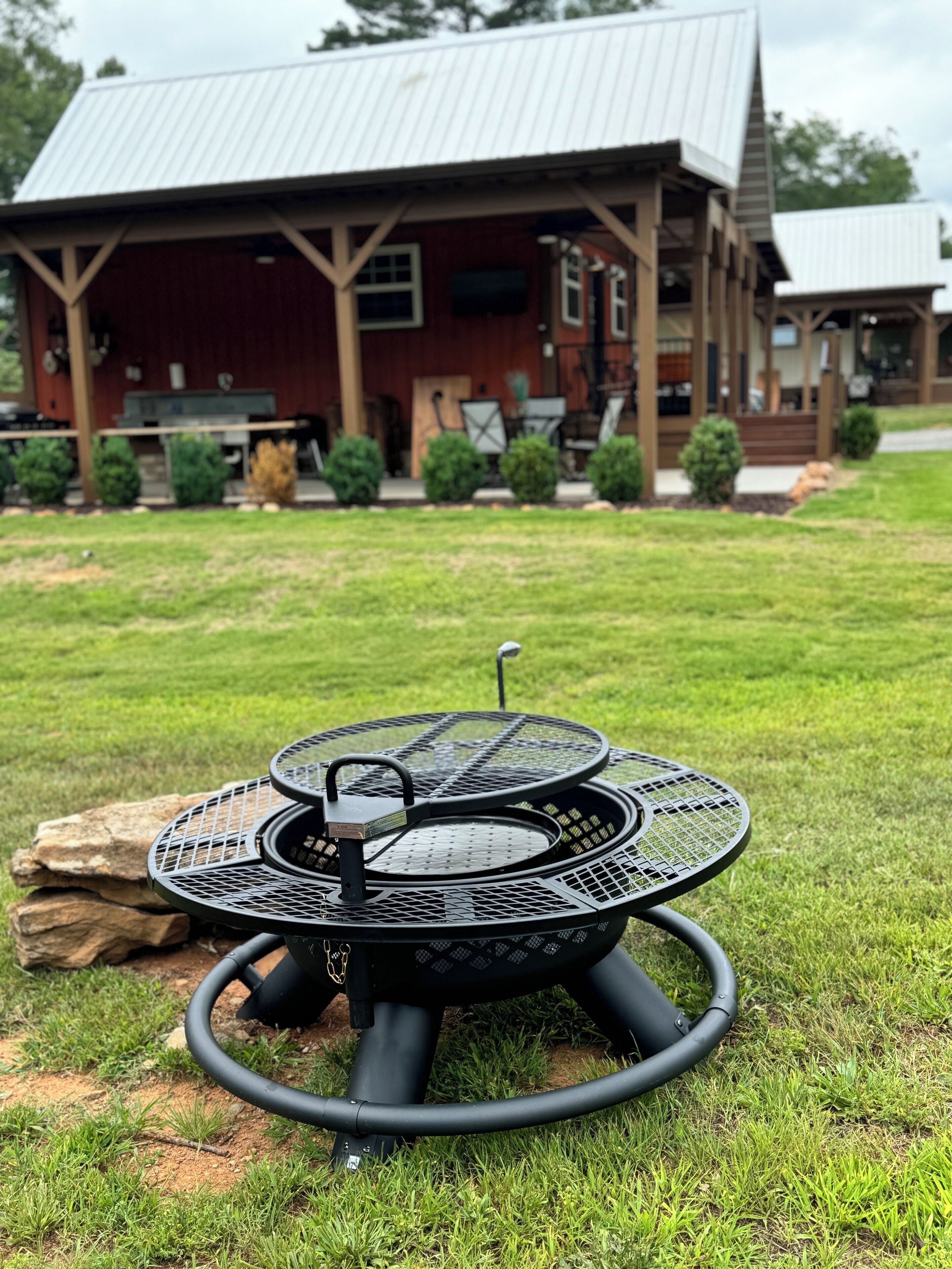 A metal outdoor grill with a circular design, placed on a grassy lawn in front of a rustic wooden building with a metal roof.