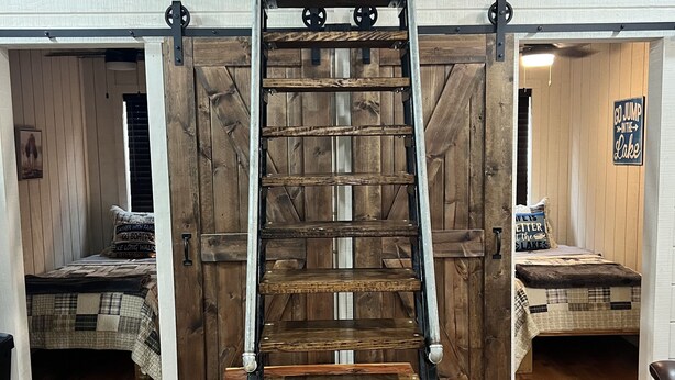 Une pièce de style « loft » avec une échelle en bois menant à une mezzanine. On y trouve deux lits avec une literie à carreaux, un plancher en bois et un panneau indiquant : « Allez, sautez dans le lac ! »