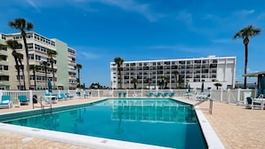 Una piscina rettangolare con bordo bianco, circondata da sdraio e palme, con un edificio di un hotel sullo sfondo.