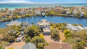 A coastal residential area with houses along a water body, a clear sky, and a prominent blue circular sign. - Only Good Vibes|Golf Cart to Beach|Lakefront Home w/ Stunning Views & Pool (Destin)