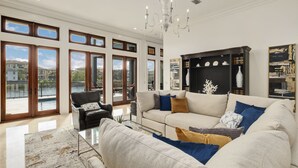 A spacious living room with a large sofa, a black armchair, and a glass coffee table. There is a chandelier hanging from the ceiling and a bookshelf against the wall. - Only Good Vibes|Golf Cart to Beach|Lakefront Home w/ Stunning Views & Pool (Destin)