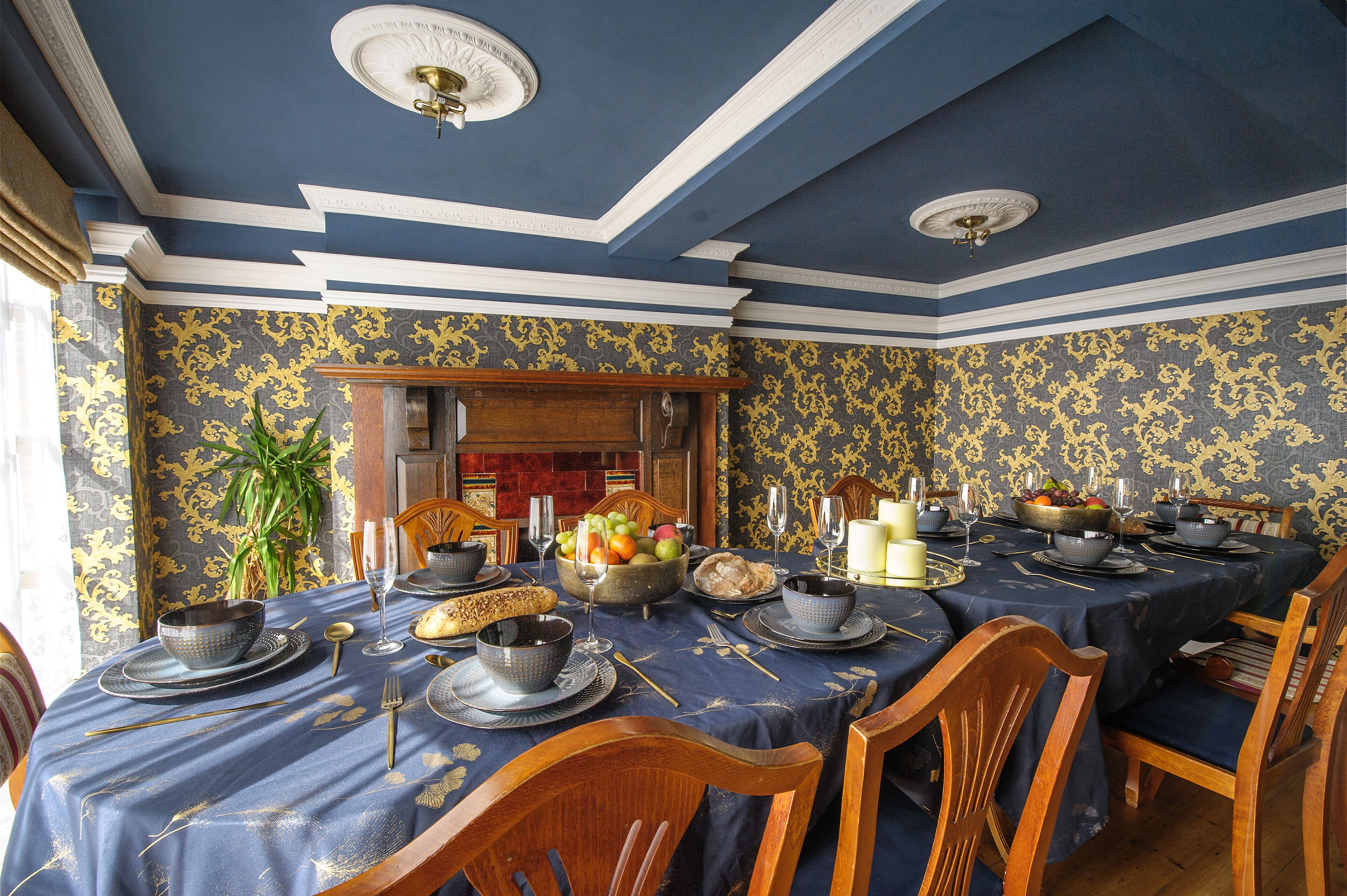 Un comedor formal con papel tapiz azul y dorado, sillas de madera y una mesa dispuesta para una comida.