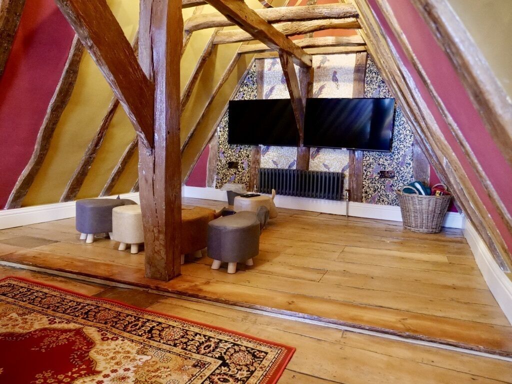 Habitación de desván con vigas de madera a la vista, un televisor de pantalla plana en la pared, una alfombra estampada y un rincón de estar con otomanas.