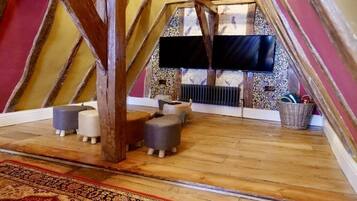 Habitación de desván con vigas de madera a la vista, un televisor de pantalla plana en la pared, una alfombra estampada y un rincón de estar con otomanas.