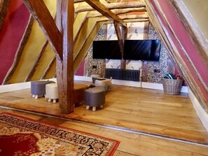 Habitación de desván con vigas de madera a la vista, un televisor de pantalla plana en la pared, una alfombra estampada y un rincón de estar con otomanas.